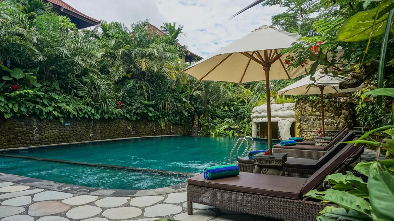 A turquoise pool with stone edging and a stone-tiled deck has sun loungers, cream umbrellas and rolled towels beside it, set against dense tropical greenery.