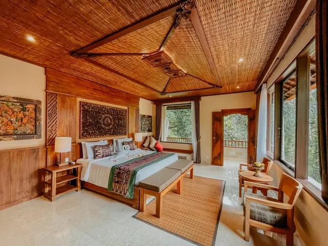 A bedroom with a king bed, a woven ceiling and wooden furnishings, with large windows and an open door onto a balcony overlooking trees.
