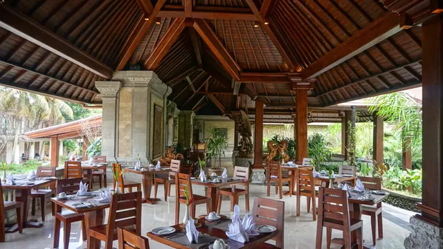 An open-sided Balinese-style restaurant with wooden tables and chairs set with folded napkins, a timber roof and carved stone pillars, with palm trees visible outside.
