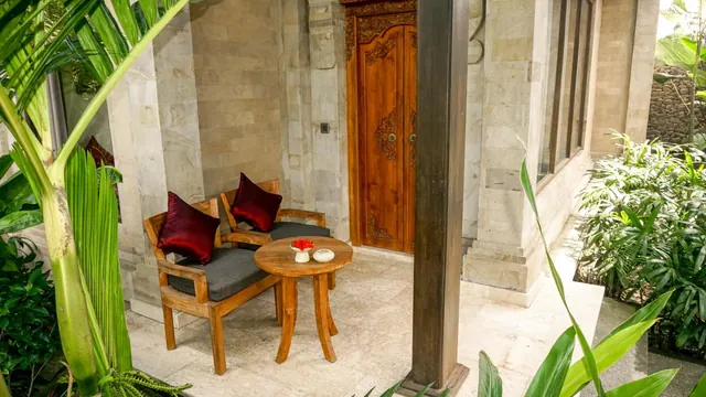 A covered stone terrace with two wooden armchairs with grey cushions and red pillows, a small round wooden table with flowers, and a carved wooden door, surrounded by tropical plants.