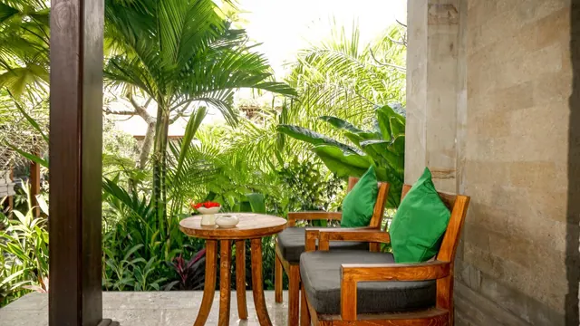 A covered terrace with two wooden armchairs with green cushions and a round wooden side table with red flowers, looking out onto lush tropical garden.