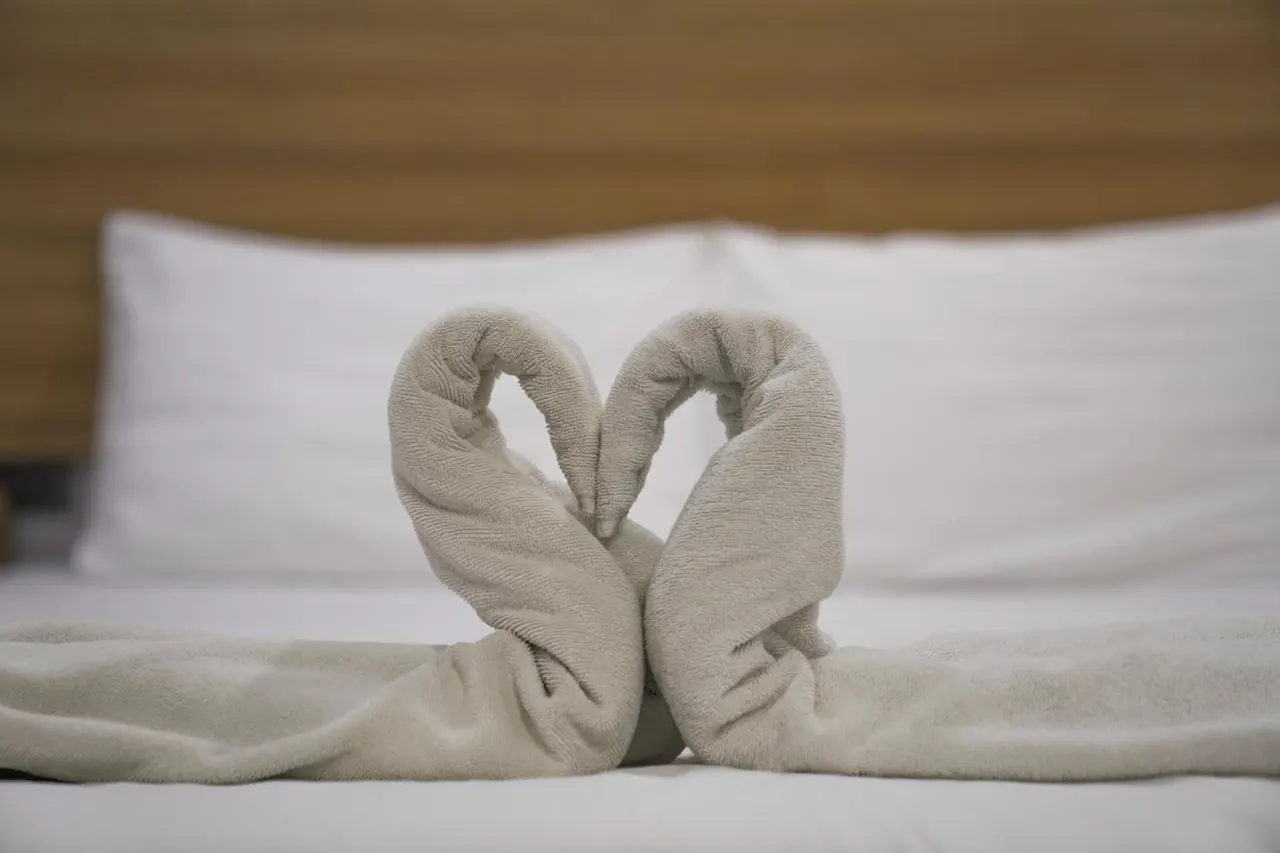Two towels folded into swan shapes forming a heart rest on a made bed in front of white pillows and a wooden headboard.