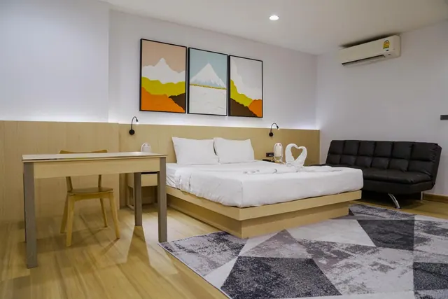 A staged bedroom with a king bed dressed in white linen, three abstract mountain paintings, a wooden desk and chair, a black sofa and a wall-mounted air conditioner.