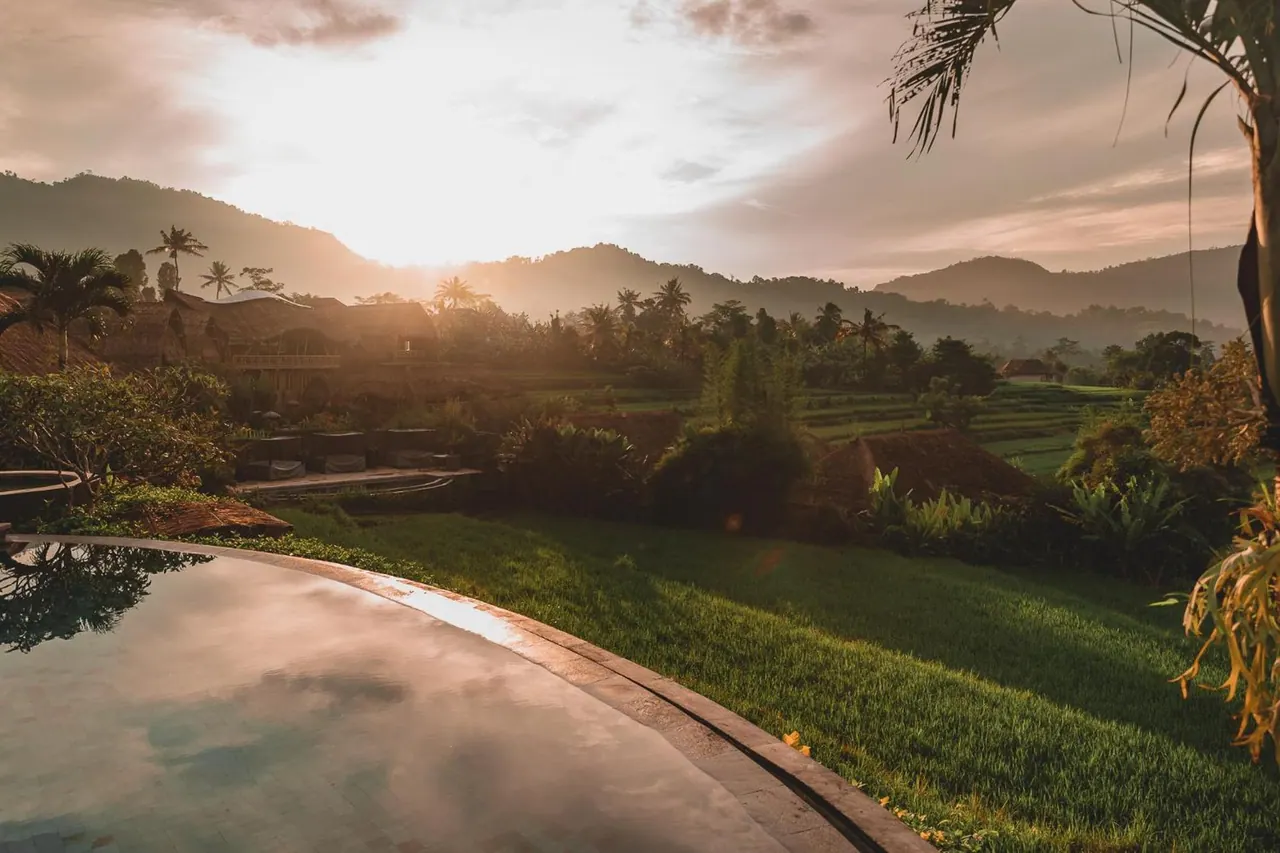 Sunset over misty mountains and rice terraces, seen from the curved stone edge of a pool with grass and palm trees nearby.