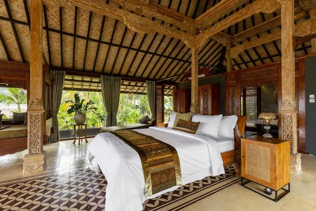 A bedroom with a king bed, carved wooden pillars and a thatched ceiling, with large windows opening onto a tropical garden.