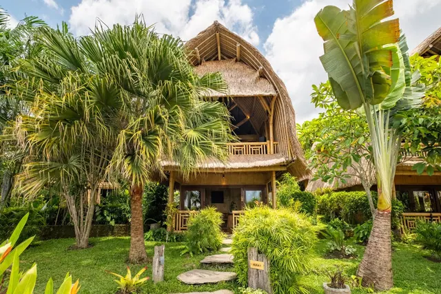 A two-storey thatched bamboo villa stands among palm trees and a lawn with a stepping-stone path.