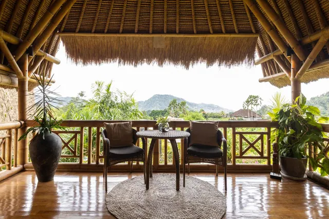 A bamboo-roofed balcony with two wicker chairs, a round table and potted plants, overlooking green hills and trees.