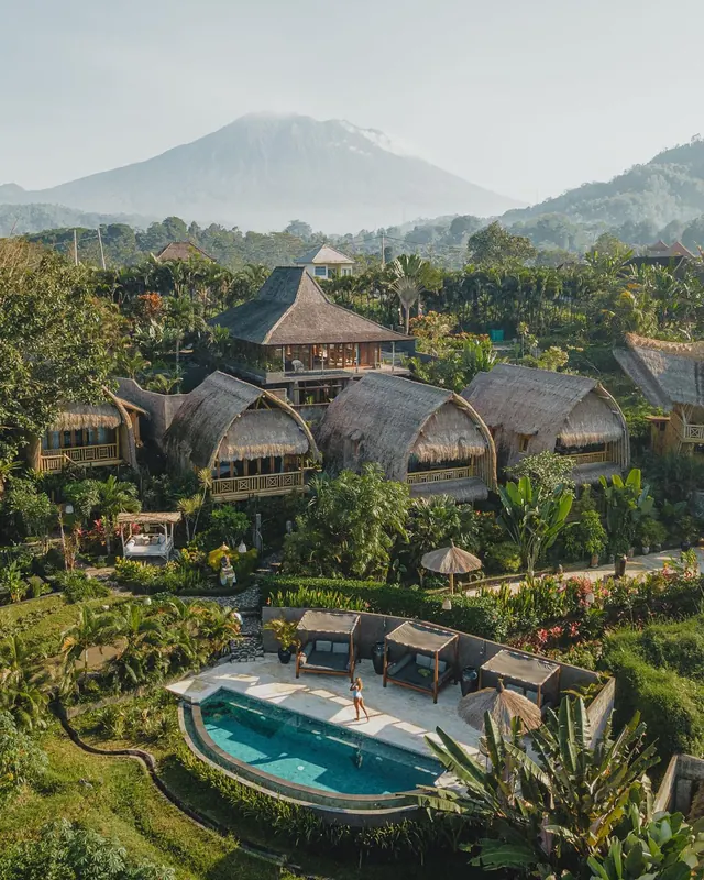 Aerial view of a resort with thatched bungalows, a small swimming pool with sun loungers, and a volcano in the background.