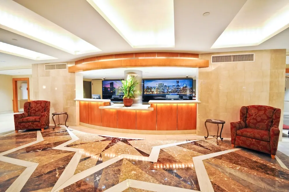 Wide view of a hotel lobby with a curved wood reception desk, two red armchairs, a potted plant and a patterned marble floor.