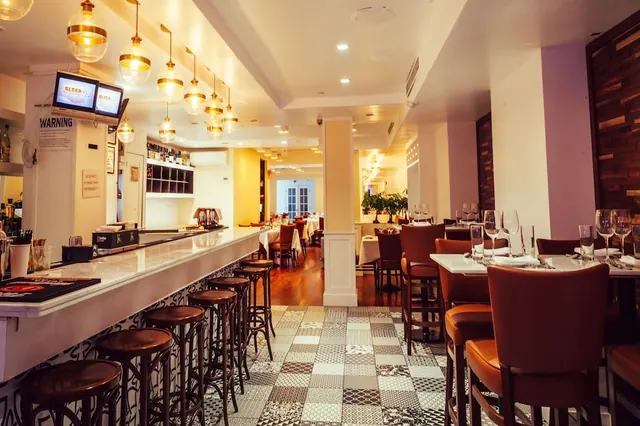 A restaurant dining room with a long bar counter, bar stools, pendant lights and tables set with wine glasses.