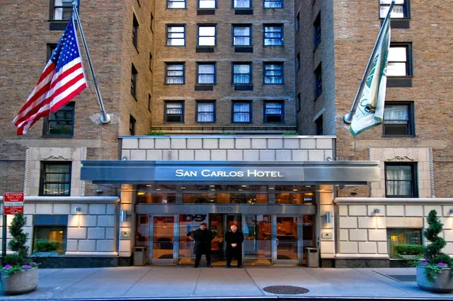 The brick front of the San Carlos Hotel with a metal canopy over the glass entrance doors, an American flag and a green and white flag on poles, and two men standing at the doors.