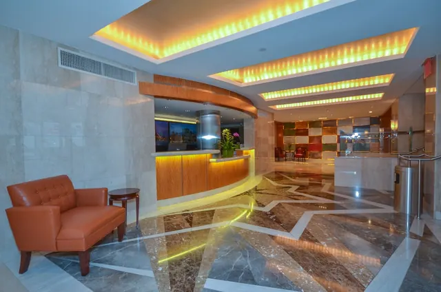 A hotel lobby with a curved wooden reception desk, a tan leather armchair, a marble floor with inlaid patterns, and backlit yellow ceiling coves.