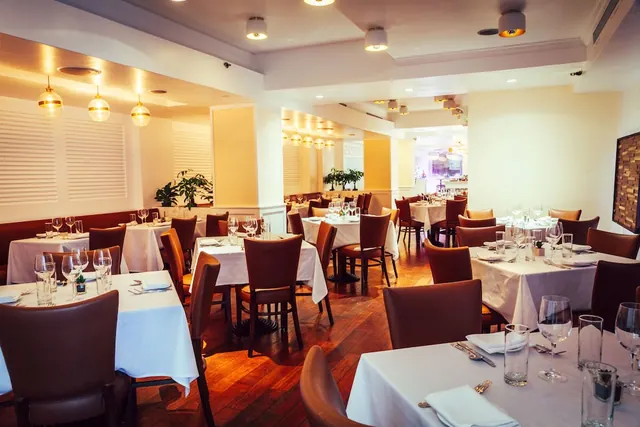 A dining room with white-clothed tables, brown upholstered chairs, pendant lights and a wooden floor.