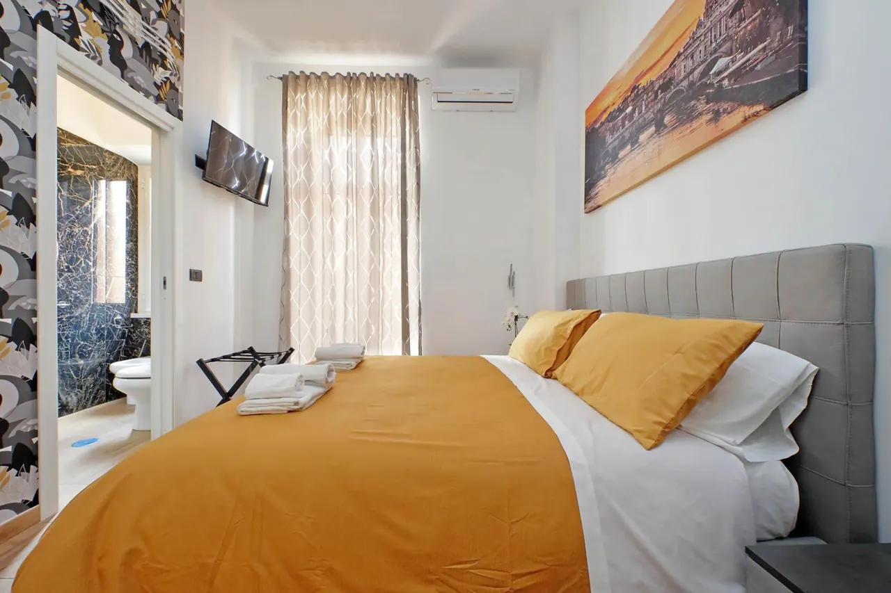 A double bed with an orange throw and grey upholstered headboard sits in a bedroom with a wall-mounted television and an air conditioner, and a marble-walled bathroom is visible through the open doorway on the left.