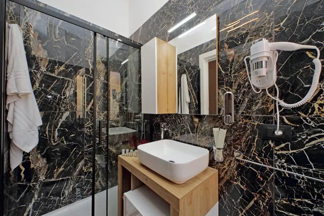 A bathroom with black and gold veined marble walls, a glass-enclosed walk-in shower, a wooden vanity with a white basin, and a wall-mounted hair dryer.