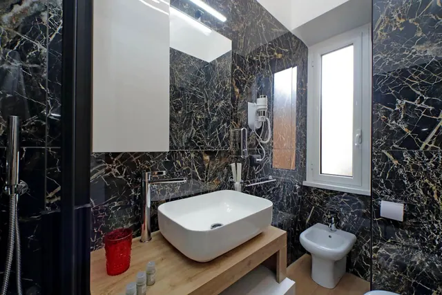 A bathroom with black marble walls, a white vessel basin on a wooden vanity, a bidet, a handheld shower and a frosted window.