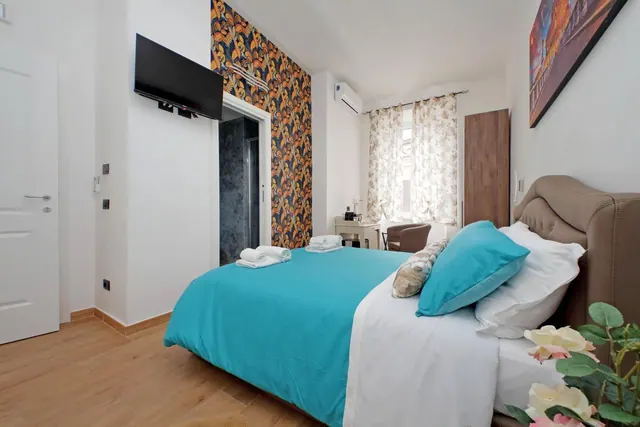 A bedroom with a teal-covered double bed, a wall-mounted television, a patterned accent wall, a desk by the window, and a wooden wardrobe.