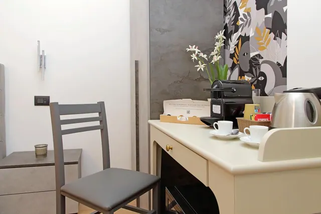 A cream desk in a hotel room holds a coffee machine, a steel kettle, two cups on saucers, a tea basket and a white orchid, beside a grey upholstered chair.