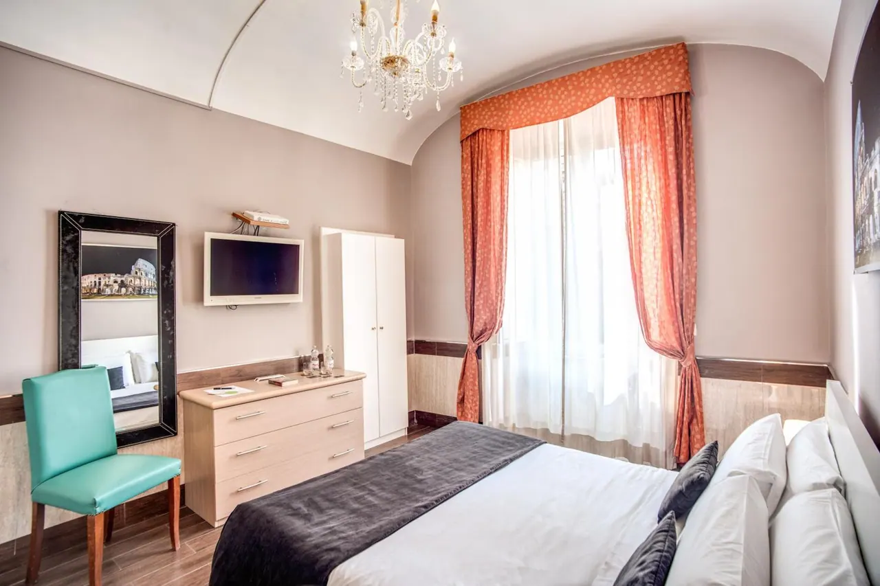A bedroom with a double bed under a vaulted ceiling and chandelier, a wall-mounted television, a dresser, a white wardrobe, and a mirror reflecting a picture of the Colosseum.