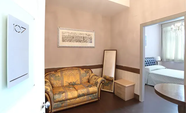 A sitting area with a floral sofa, a framed print of Rome, a floor mirror and a nightstand, with an adjoining bedroom visible through an open doorway.