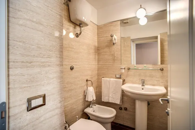 A bathroom with travertine-style wall tiles, a pedestal sink, a bidet, a toilet, hanging towels and a wall-mounted cistern.