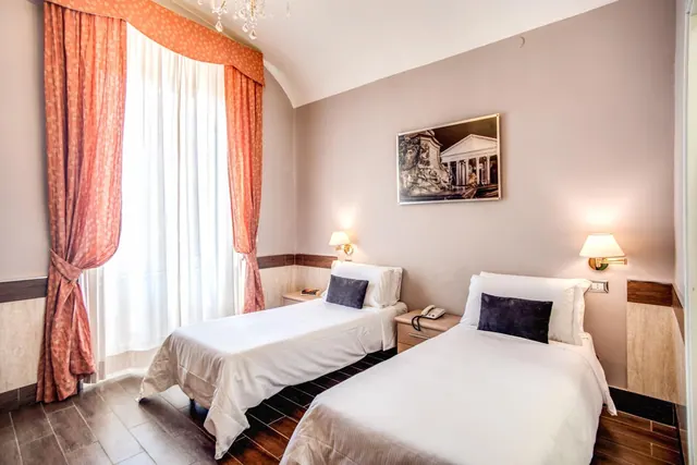 A twin bedroom with two single beds with white bedding, wall lamps, a framed print of the Pantheon, and orange curtains beside a window.