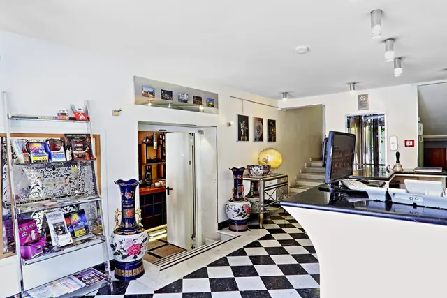 A hotel reception area with a black and white checkered marble floor, a reception desk with computer monitors, a brochure rack, large decorative vases and a staircase.
