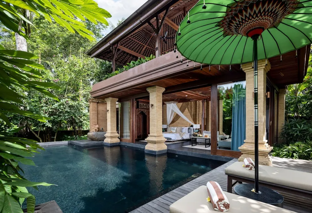 A private pool sits beside an open-sided villa bedroom with a canopy bed, sun loungers and a green parasol, surrounded by tropical greenery.