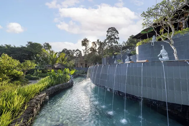 A stone-edged water channel runs beside lush tropical plants, with water spilling over a tiled wall topped by white statues.