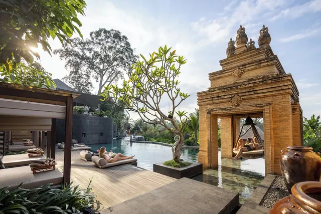 Two people lie on sun loungers beside a pool, with a carved stone gateway and a hanging daybed to the right.