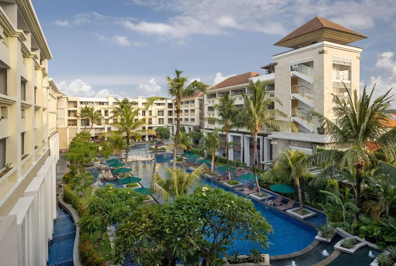Elevated view of a winding resort swimming pool with sun loungers under green umbrellas, surrounded by palm trees and multi-storey hotel buildings.