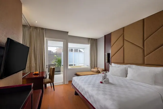 A bedroom with a king bed, a wall-mounted television, a wooden desk and chair, and a glass door opening onto a terrace with plants.