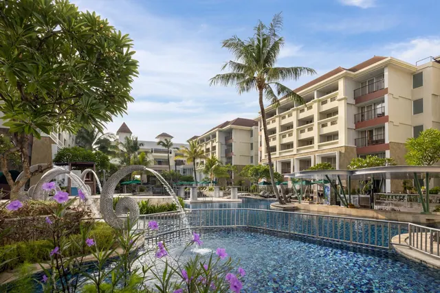 A large swimming pool with a sculptural water fountain and purple flowers in the foreground, with a palm tree, a covered bar and a multi-storey hotel building behind.