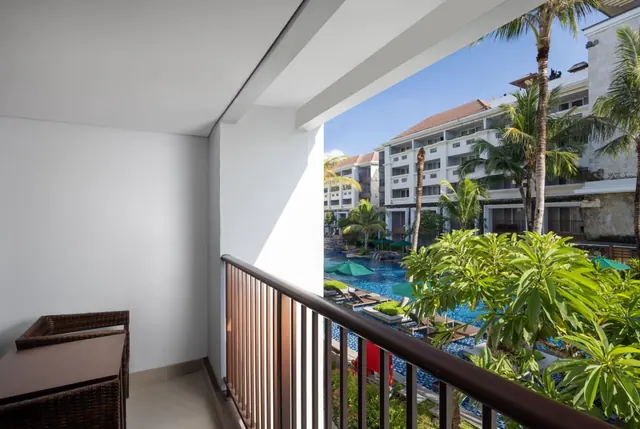 A balcony with wicker furniture overlooks a swimming pool with green umbrellas, sun loungers and palm trees beside hotel buildings.
