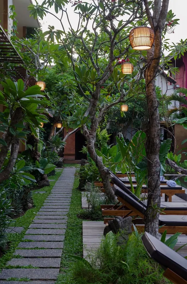 A stone-paved path runs between grass and frangipani trees in a garden, with woven lanterns hanging from the branches and sun loungers on a timber deck at right.