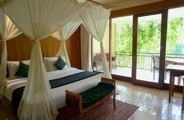 A bedroom with a canopy bed under a mosquito net and a bench at its foot, with glass doors opening onto a balcony with chairs and a view of trees.