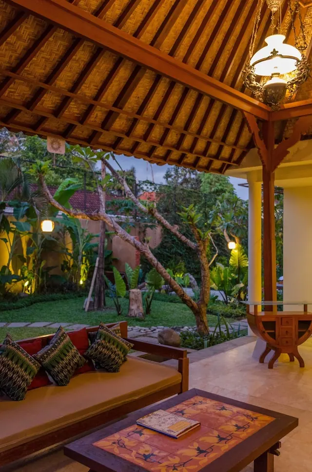 A covered open-sided sitting area with a wooden daybed, patterned cushions and a tiled coffee table, looking onto a lawn and garden at dusk.
