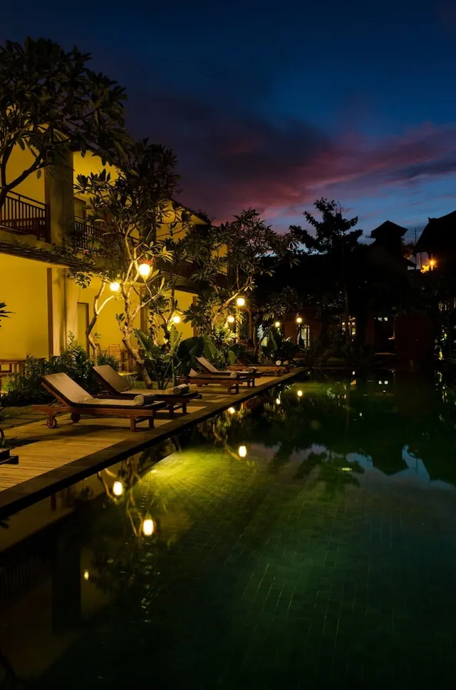 Illuminated pool at dusk with wooden sun loungers along the deck, frangipani trees and a yellow building beside it.