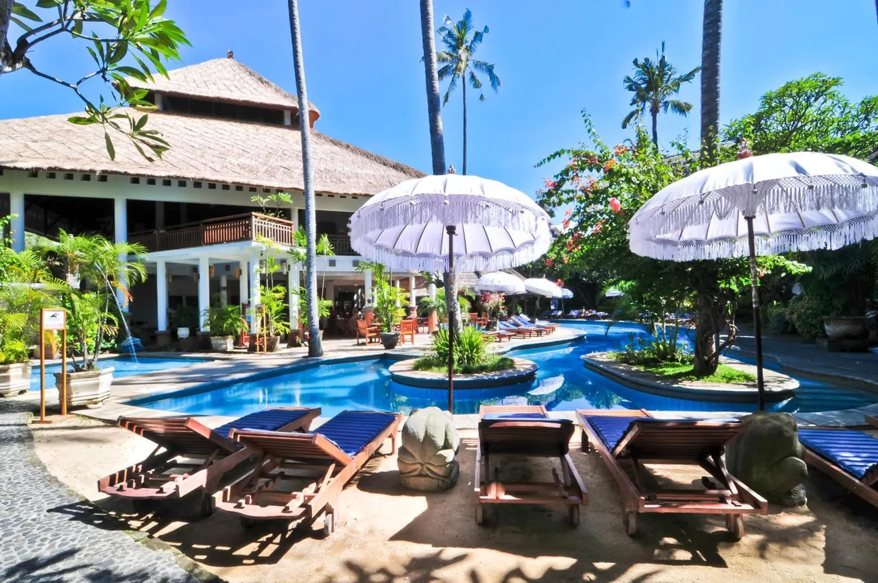 A large swimming pool with white fringed umbrellas and wooden sun loungers, with a thatched-roof building and palm trees behind it.
