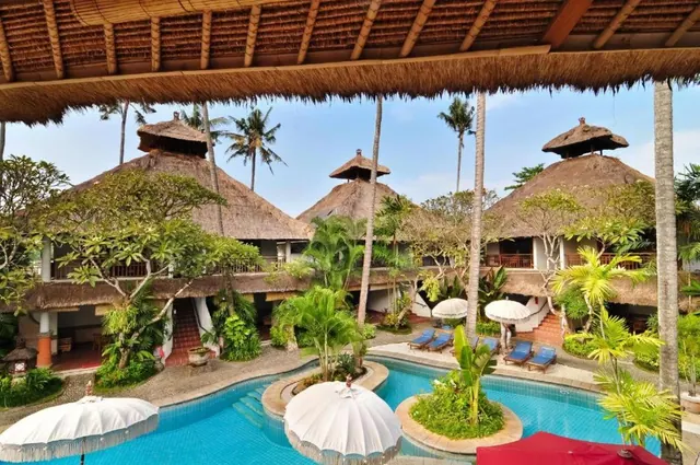 A curved swimming pool with small planted islands, white umbrellas and sun loungers sits in front of thatched-roof bungalows, photographed from under a thatched pavilion.