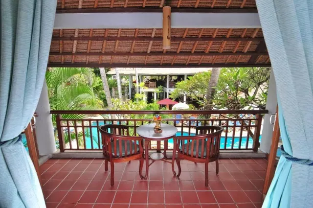 A tiled balcony with two wooden chairs and a round table overlooks a swimming pool and palm trees, framed by blue curtains under a thatched roof.