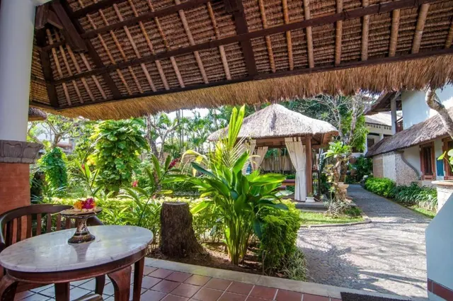 A covered thatched terrace with a round marble table and wooden chair overlooks a garden with a thatched gazebo and a stone path.