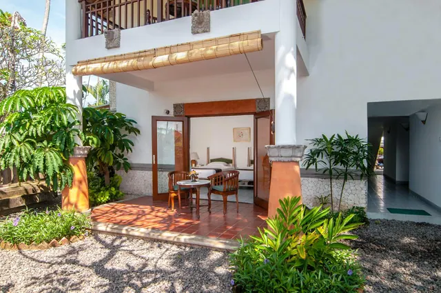 The exterior of a ground-floor room shows a tiled terrace with two wooden chairs and a small round table, open glass doors leading to a bed inside, and a bamboo blind above.