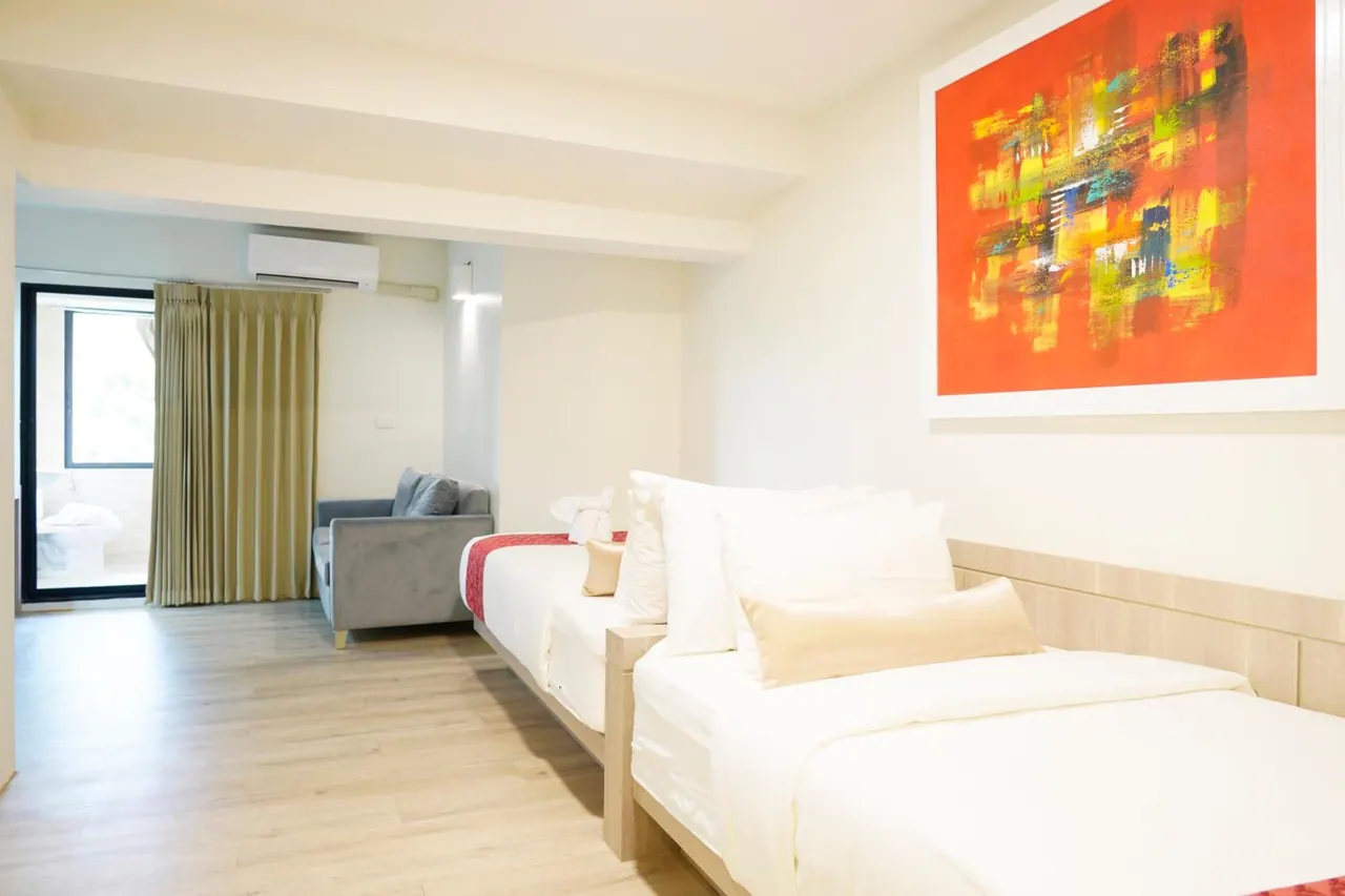 A bedroom with two twin beds, a grey sofa, a wall-mounted air conditioner and a red abstract painting, with a glass door leading to a balcony.