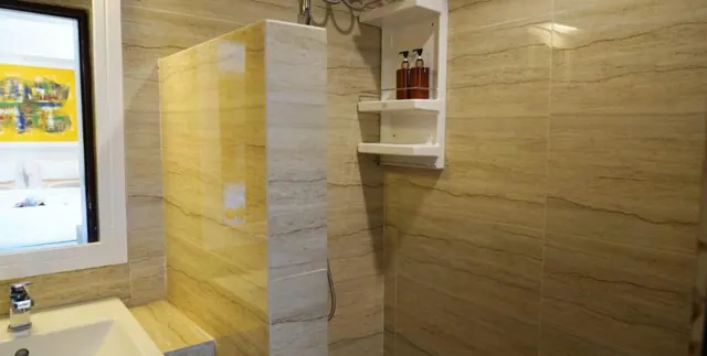 A bathroom with veined stone-look wall tiles, a walk-in shower area with a handheld shower hose, a white corner shelf holding two amber bottles, and a white sink at the lower left.