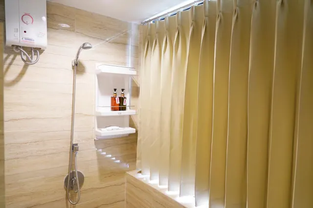 A tiled shower with a handheld shower head, a wall-mounted water heater, a corner shelf holding two bottles, and a beige shower curtain on a raised ledge.