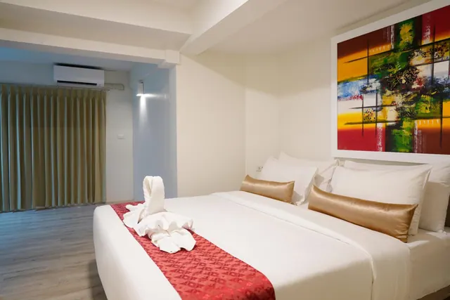 A bedroom with a made-up double bed with white linens, a red runner, a towel swan, gold bolster pillows, a large abstract painting on the wall, and a wall-mounted air conditioner.