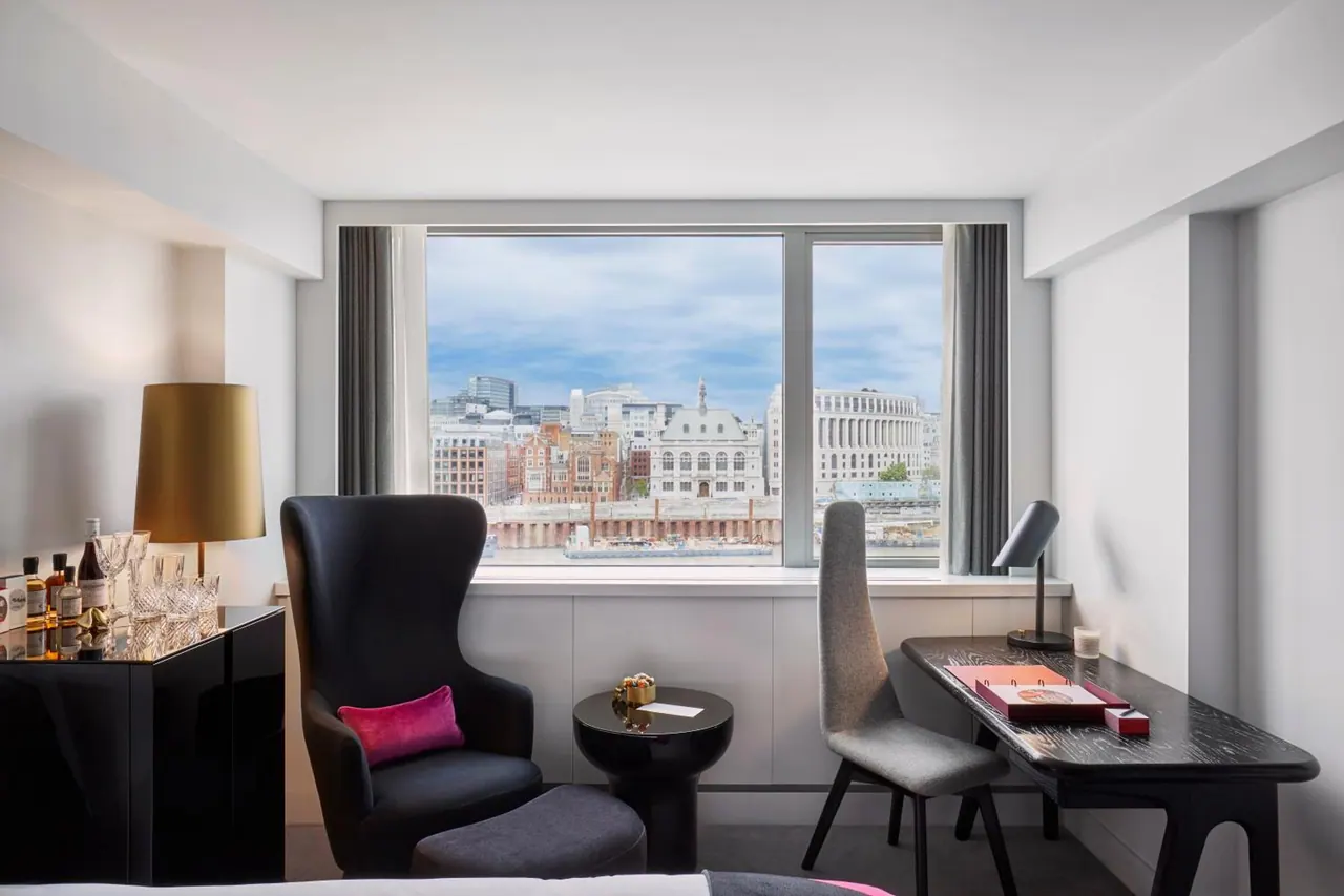 A hotel room with an armchair, a small round side table and a desk with a chair beside a large window looking over city buildings.