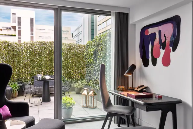A hotel room with a desk, lamp and armchair beside sliding glass doors that open onto a terrace with a table, chairs and planters.
