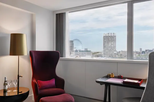 A burgundy velvet armchair with a footstool, a brass floor lamp and a desk sit beside a window looking out over the London Eye and a tall white office tower.